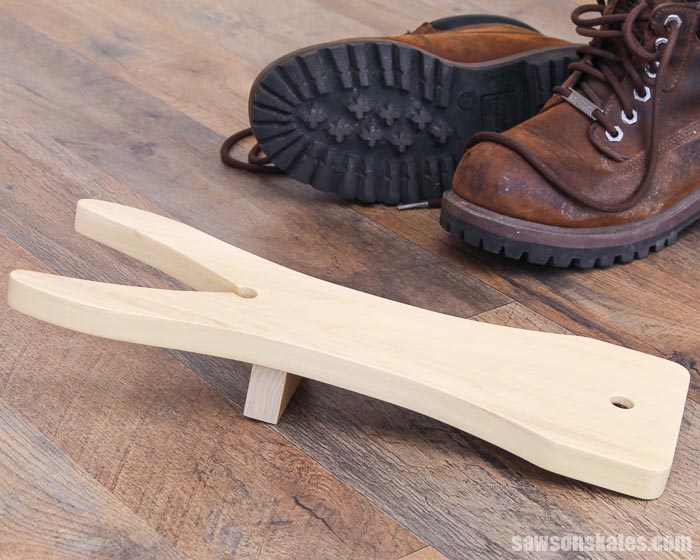 Side view of a wooden DIY boot jack in the foreground and a pair of boots in the background