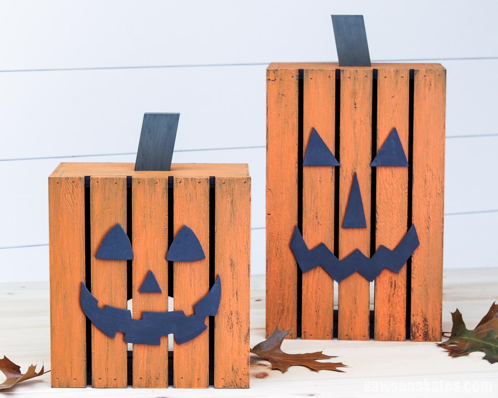 A short and tall wooden DIY jack-o-lantern with several leaves on a table