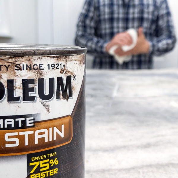 Can of wood stain in the foreground with person wiping their hands in the background