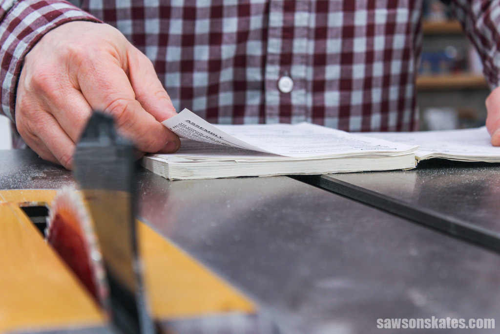 Table saw blade and riving knife in the foreground with a hand turning the page of a manual in the background