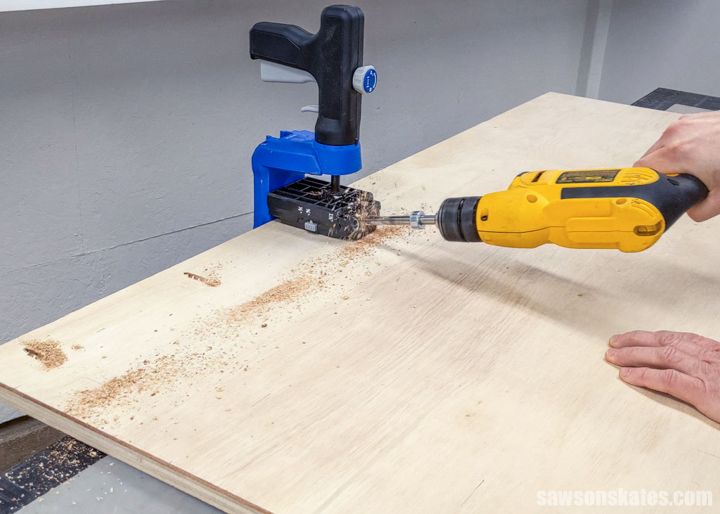 Using a drill and a Kreg Jig to make pocket holes in a piece of plywood for a rolling shop cart