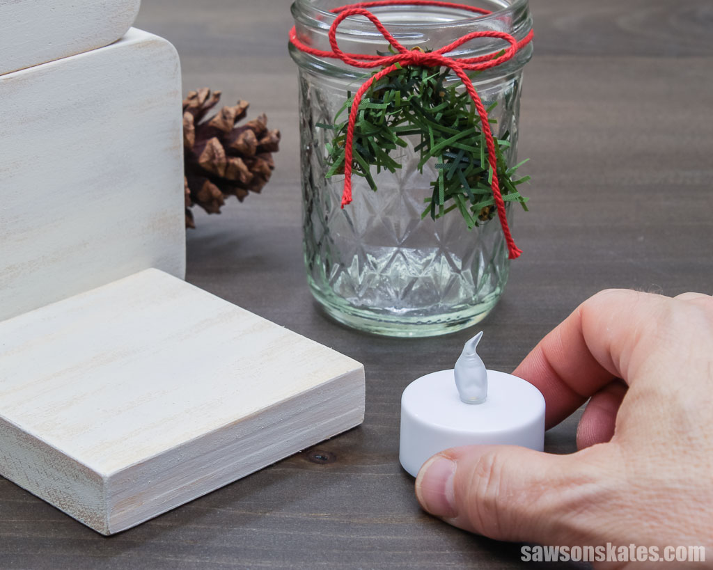 Hand picking up an LED tea light for a DIY wooden snowman with a mason jar candle holder
