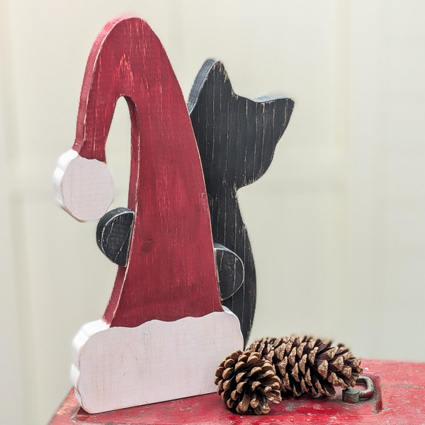 Wooden cat painted black peeking around a red Santa hat