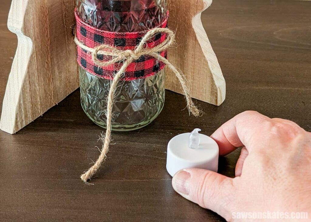 Fingers squeezing an electric tea light in front of a wood tree and mason jar wrapped with ribbon