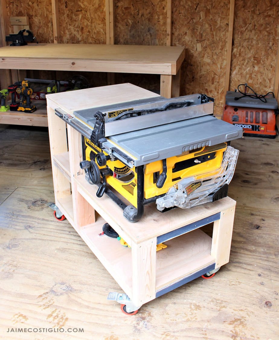 Compact DIY table saw workbench on locking casters with built-in storage, holding a portable table saw in a small workshop.