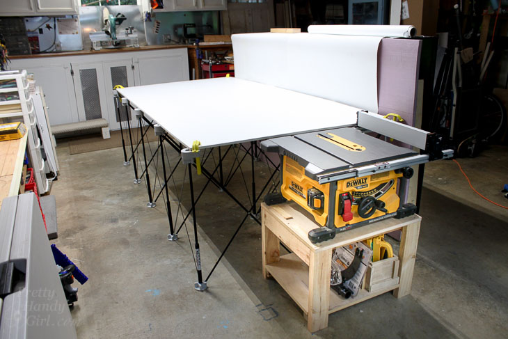 Portable table saw on a small wooden stand with a large fold-out outfeed table in a garage workshop.