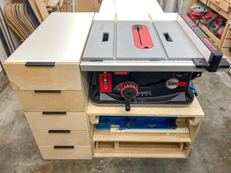 Table saw built into a wooden cabinet with side drawers and lower storage shelves in a workshop.