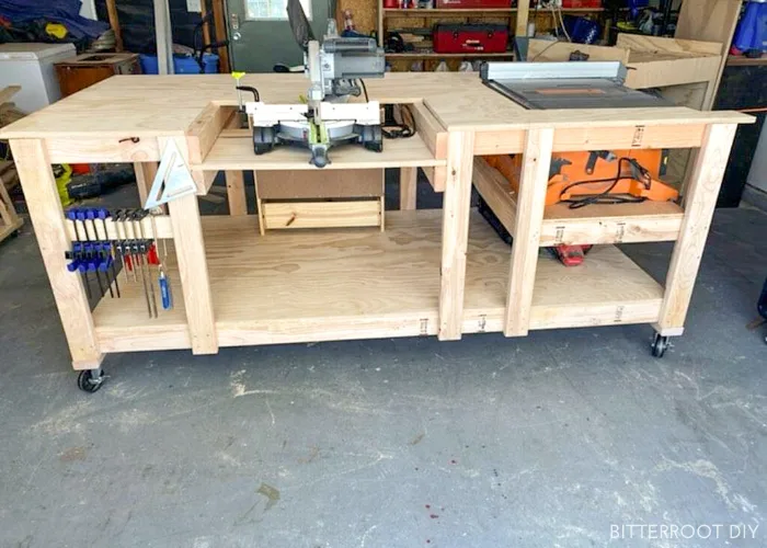 Large DIY workbench on casters with a miter saw station, built-in table saw, and storage for clamps and tools in a garage workshop.