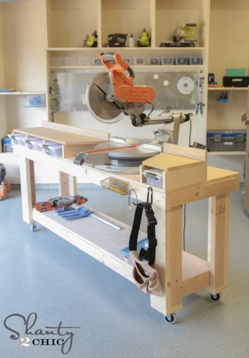 Long DIY miter saw workstation with built-in support wings, open lower storage, small side drawers, and locking casters in a garage workshop.