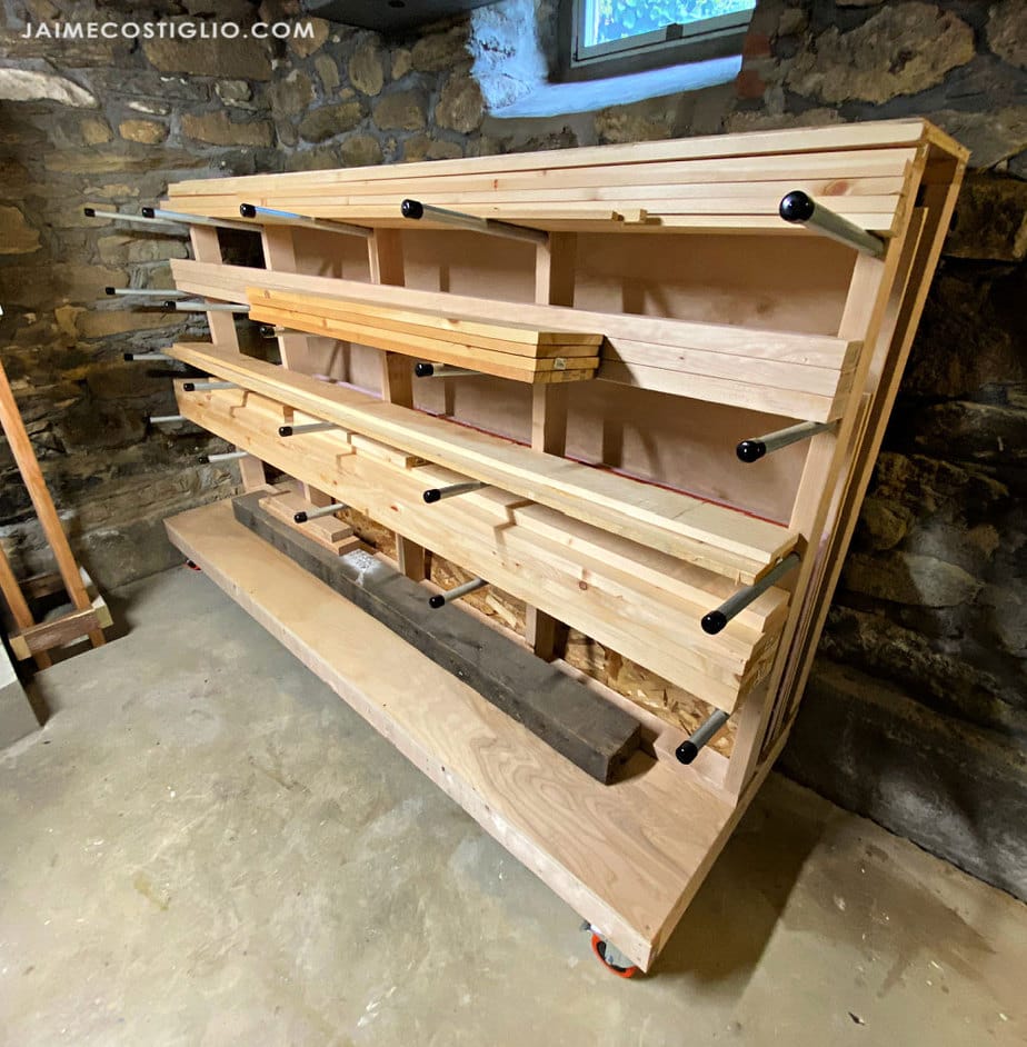 DIY rolling lumber rack with angled horizontal storage rails holding dimensional lumber, and long wood offcuts in a basement workshop.