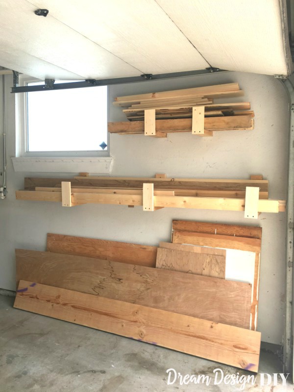 Wall-mounted scrap wood storage racks made from 2x4s in a garage, holding boards and lumber offcuts above stacked sheet goods leaning against the wall.