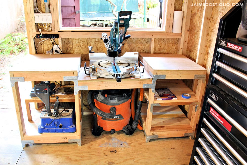 DIY miter saw station with side support tables, open shelving, metal corner brackets, and shop vac storage in a workshop.