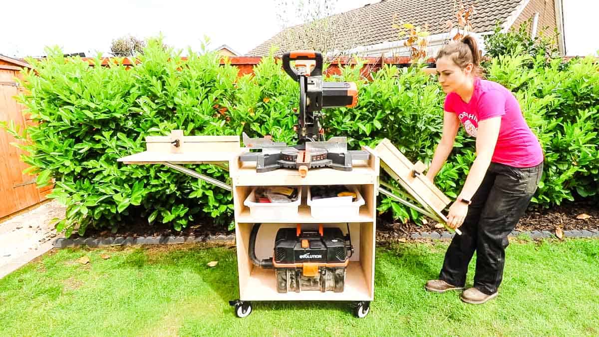 Compact mobile miter saw stand with folding support wings, built-in storage shelves, and locking casters in a yard.