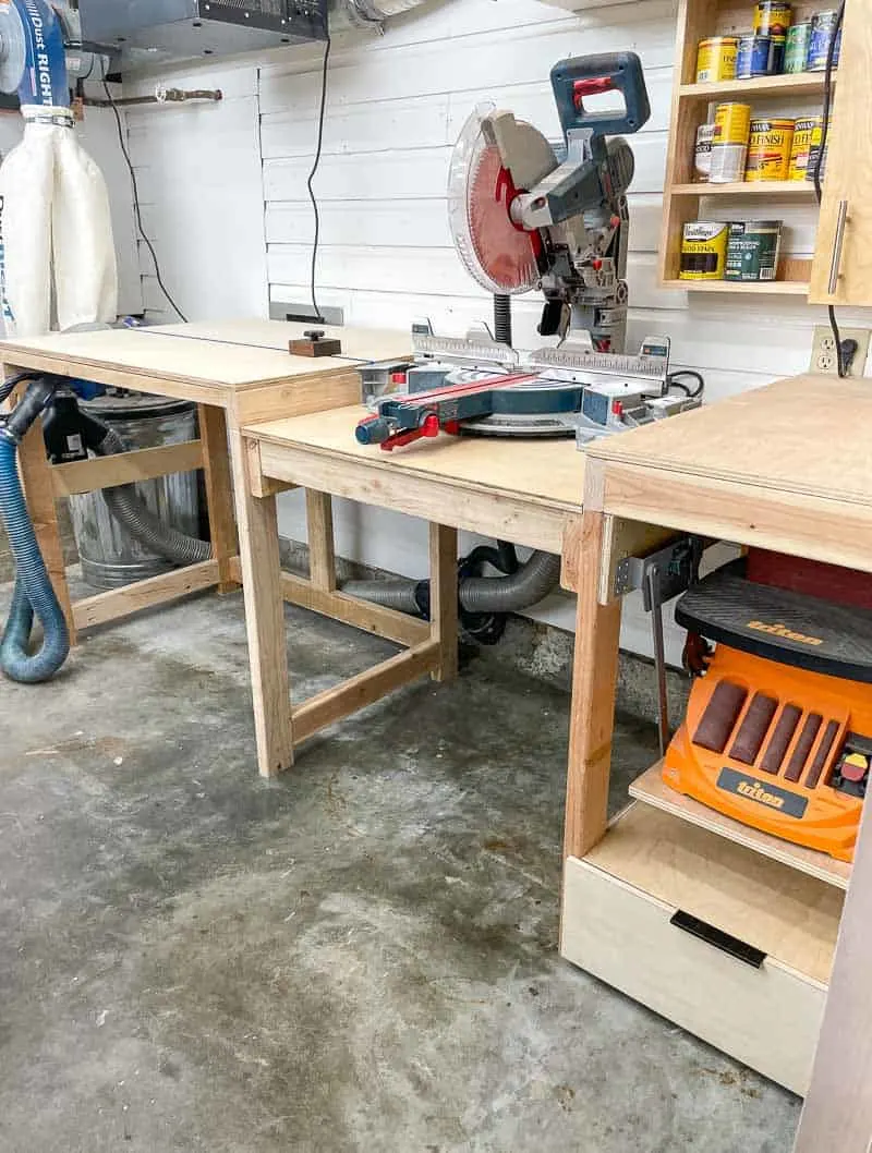 DIY miter saw workstation with built-in support tables, dust collection hose, open storage, and a lower shelf in a small garage workshop.
