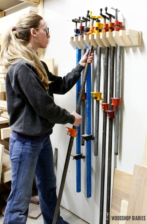Person removing a long pipe clamp from a wall-mounted wooden clamp rack in a woodworking shop.