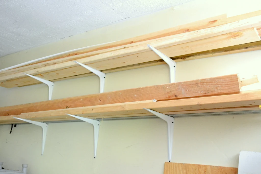 Wall-mounted lumber storage shelves made with white metal brackets and long wood boards, holding dimensional lumber near the ceiling of a garage or workshop.
