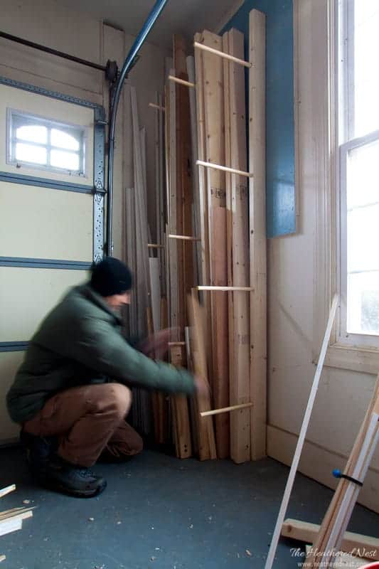 Vertical lumber storage rack built from wood dowels and side supports, holding long boards upright in the corner of a garage workshop near a window and garage door.