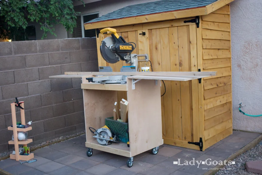 Compact rolling miter saw cart with folding side wings, built from plywood. The cart includes lower storage for tools and scrap wood and is shown next to a wooden storage shed.