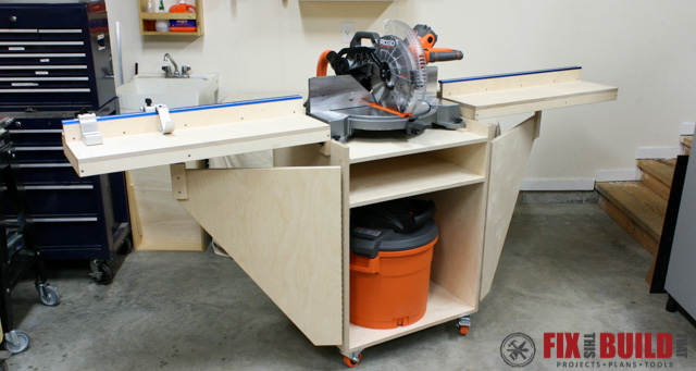 Mobile miter saw station with folding support wings, built-in shelving, and enclosed storage underneath. The workstation is mounted on casters and features long fence supports on both sides of the saw.