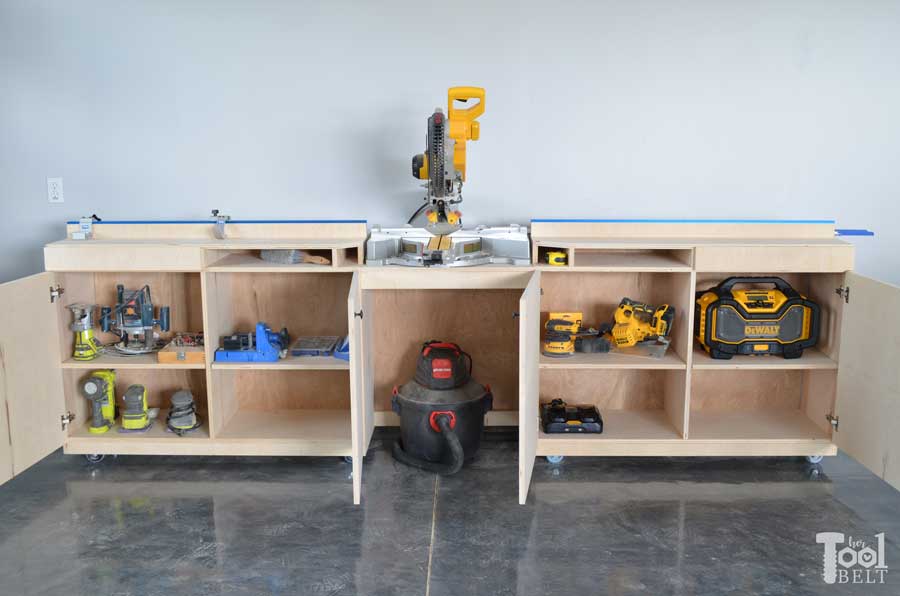 Large DIY miter saw station with built-in cabinets, adjustable fence supports, open shelving, and locking casters positioned against a gray wall.
