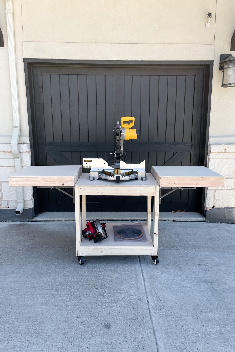 DIY mobile miter saw table with foldable support wings, lower storage shelf, and locking casters positioned in front of a black garage door.