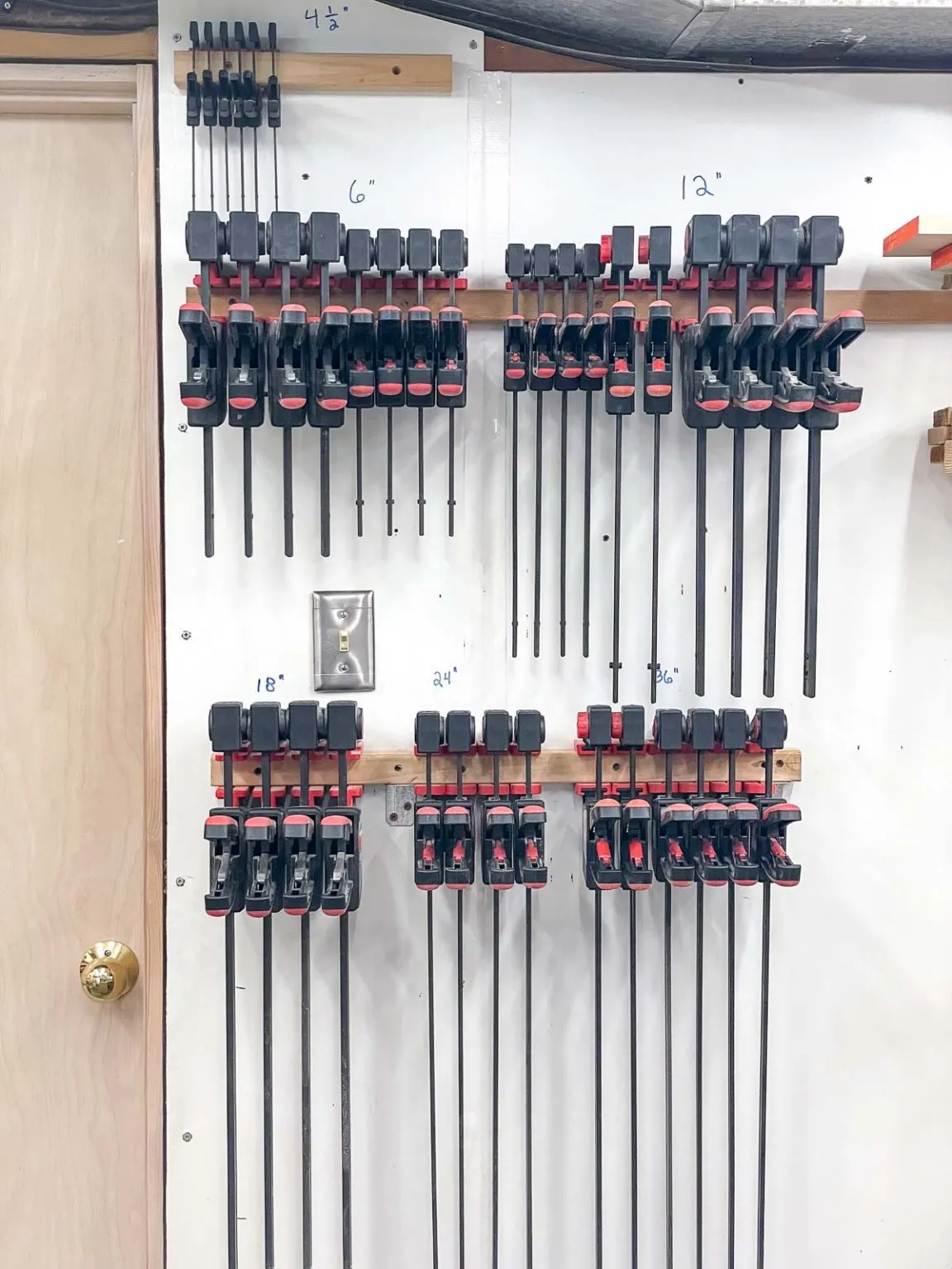 Wall-mounted bar clamp storage organized by clamp length, with multiple rows of quick-style clamps hanging from simple wooden racks in a woodworking shop.