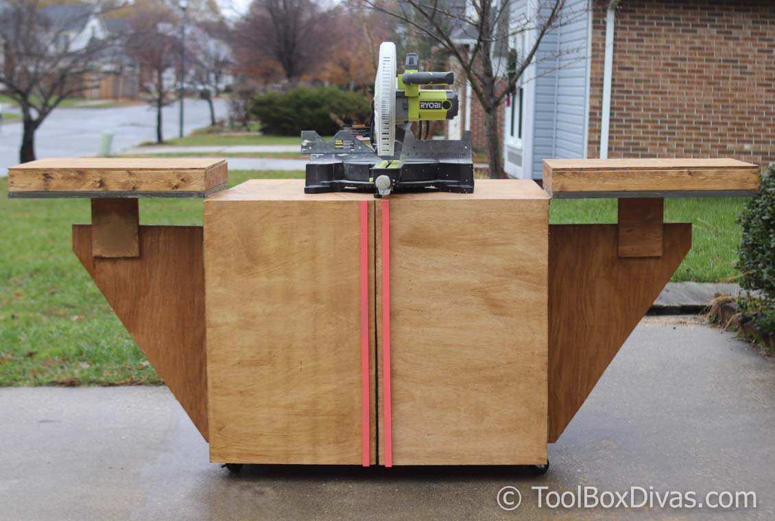Rolling DIY miter saw stand with fold-out support wings and a plywood storage cabinet mounted on casters, shown on a driveway.