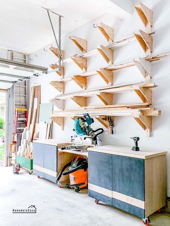 Garage workshop with a built-in miter saw station between two rolling storage cabinets beneath wall-mounted lumber racks filled with boards.