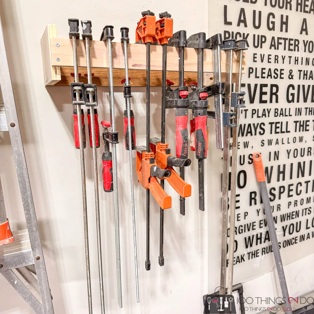 Wall-mounted DIY clamp rack with staggered wood slots for organizing bar clamps and quick-grip clamps in a small workshop.