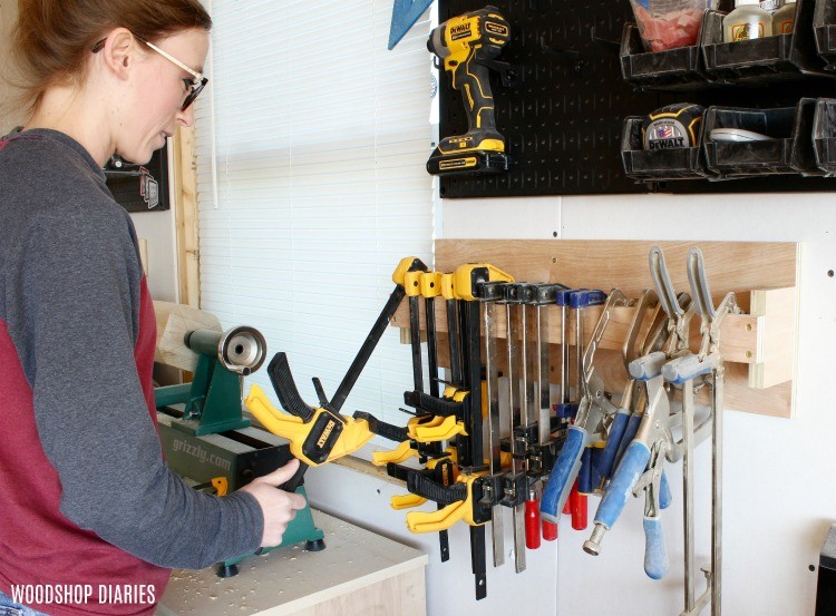 DIY wall-mounted clamp rack with organized storage for bar clamps and spring clamps beside a woodworking bench in a workshop.