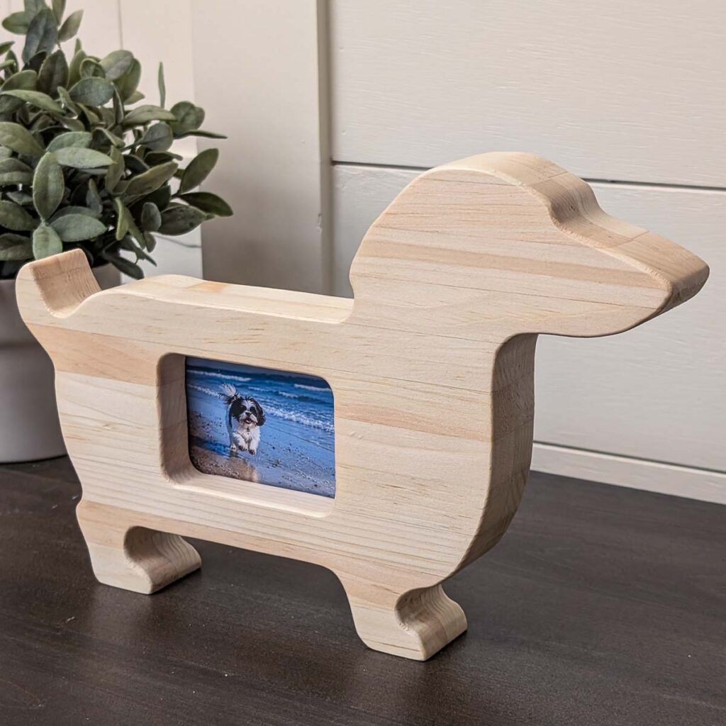 Dog-shaped wooden picture frame with a small beach photo displayed in the center, sitting on a dark tabletop beside a potted plant.