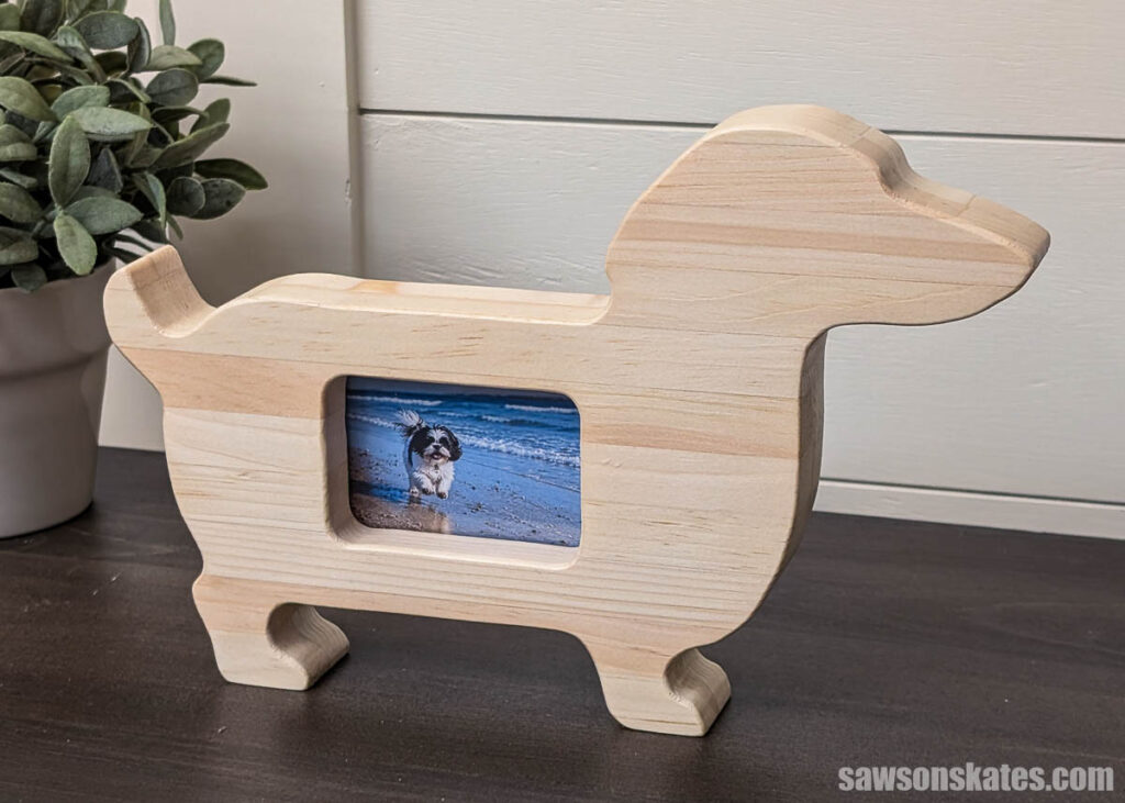 Dog-shaped wooden picture frame with a small beach photo displayed in the center, sitting on a dark tabletop beside a potted plant.