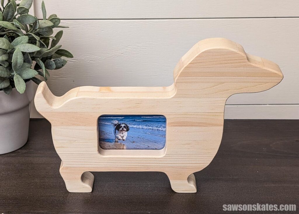 Dog-shaped wooden picture frame with a small beach photo displayed in the center, sitting on a dark tabletop beside a potted plant.