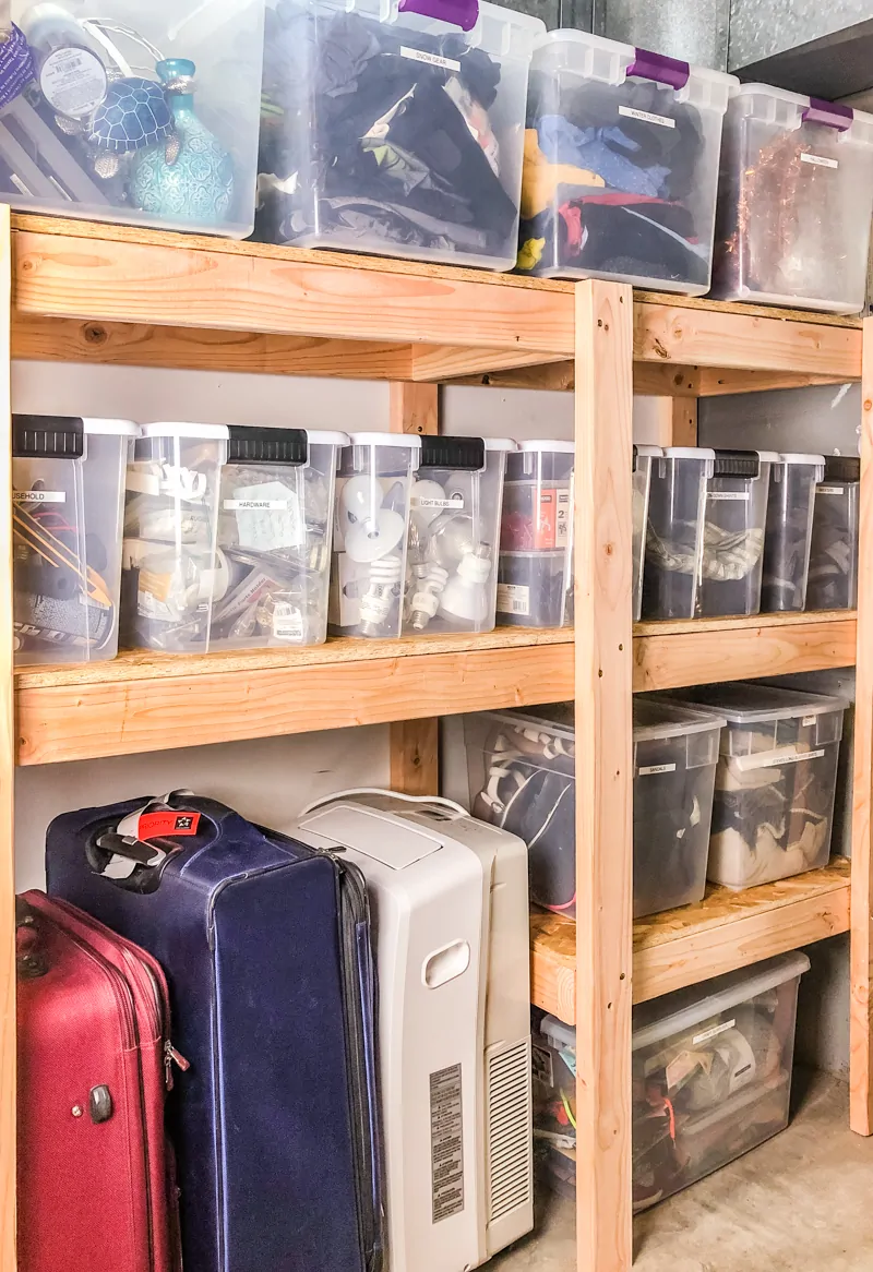 Garage storage shelves holding clear plastic bins, suitcases, and household items.