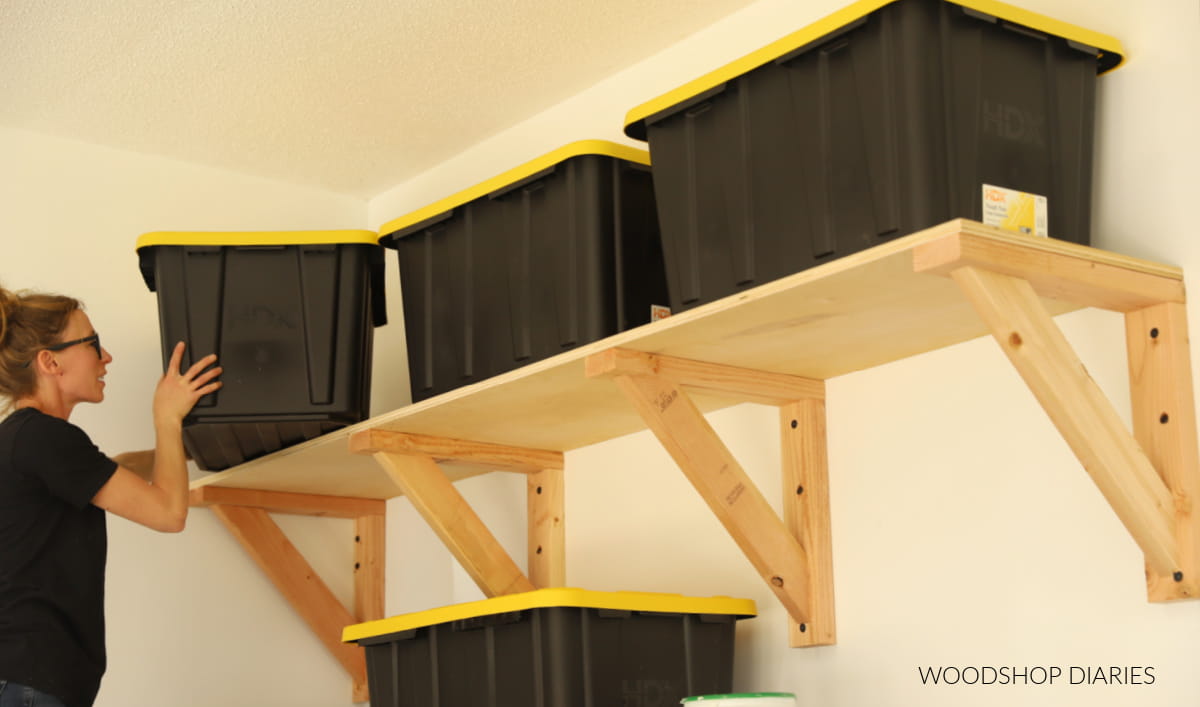 Person placing black storage totes with yellow lids on wall-mounted garage shelves supported by wooden brackets.