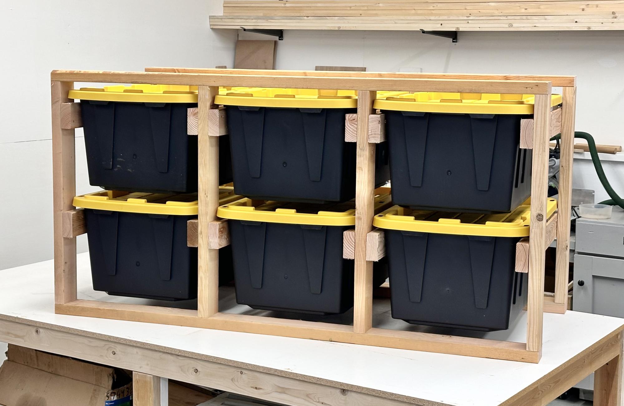 Wooden tote storage rack holding six black plastic bins with yellow lids on a workshop table.