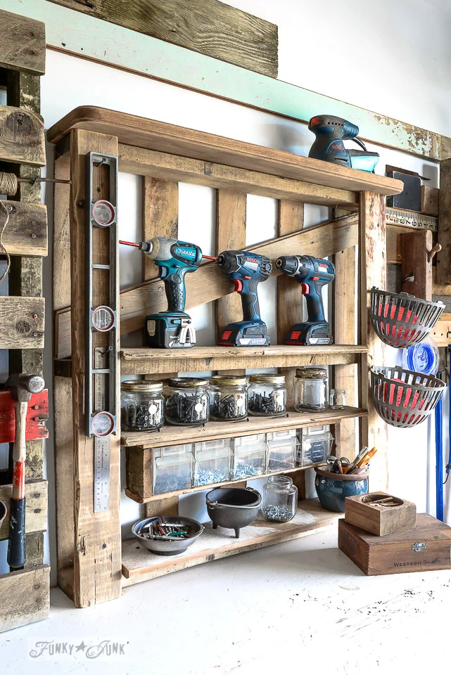 Rustic wall-mounted tool shelf holding cordless drills, jars of hardware, small bins, and hand tools in a workshop.