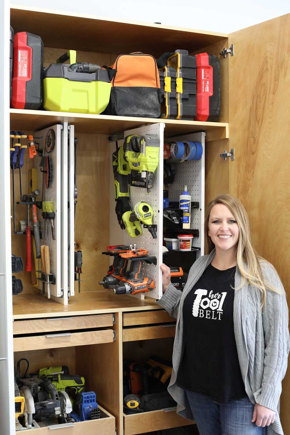 Person standing beside an open workshop storage cabinet with shelves, drawers, pegboard panels, and power tools.