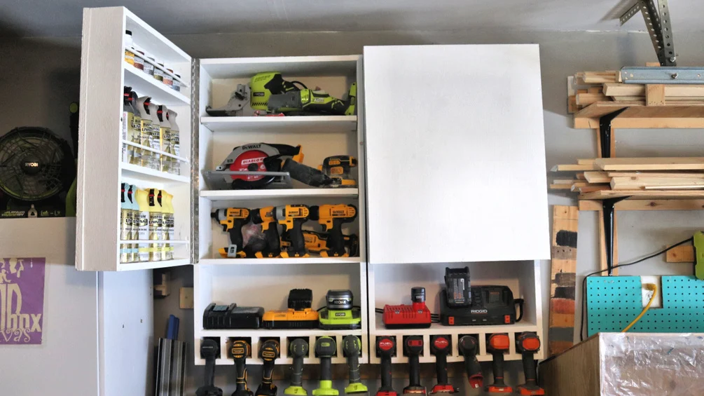 Wall-mounted workshop cabinet with open shelves holding cordless drills, sanders, batteries, chargers, and spray cans.