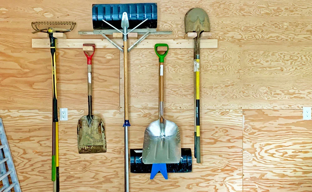 Garage wall with shovels, rakes, and snow tools hanging from a simple wood storage rack.