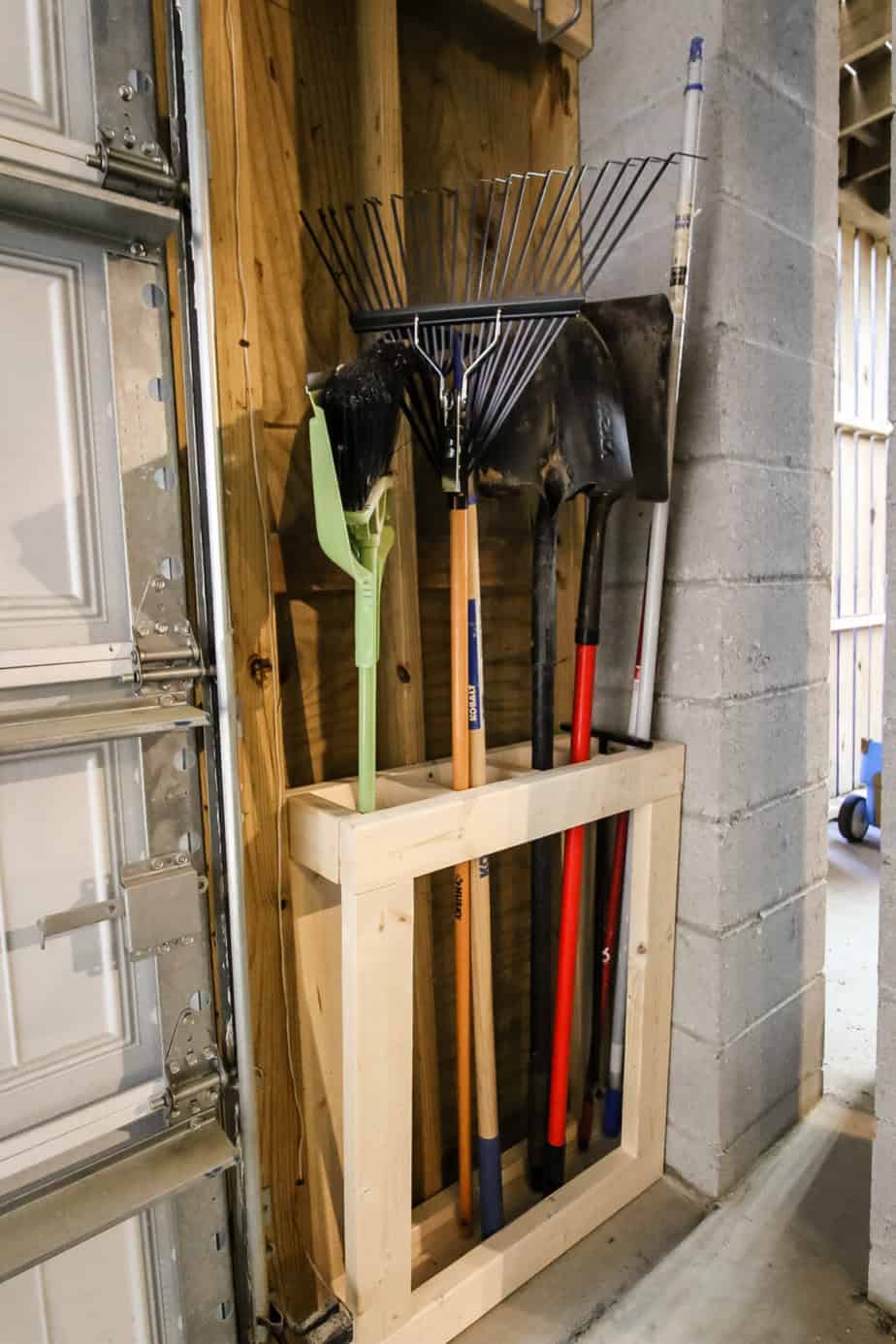 Wall-mounted garage storage rack holding rakes, shovels, and other long-handled yard tools.