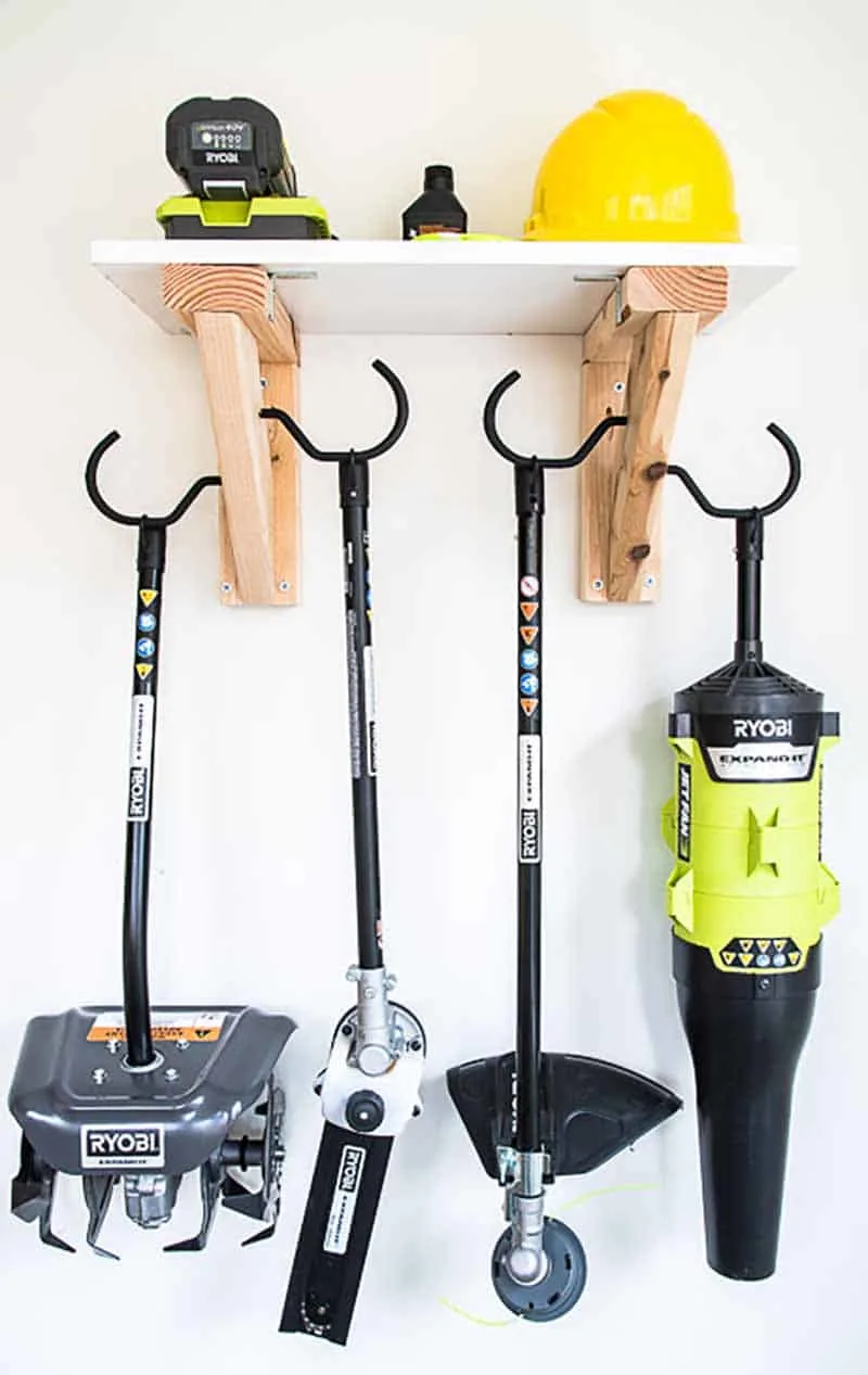 Wall-mounted garage shelf with yard tools hanging from hooks below and safety gear stored on top.
