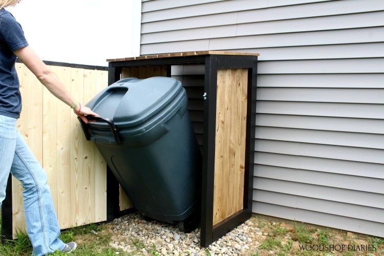 Person rolling a garbage can out from a wooden enclosure