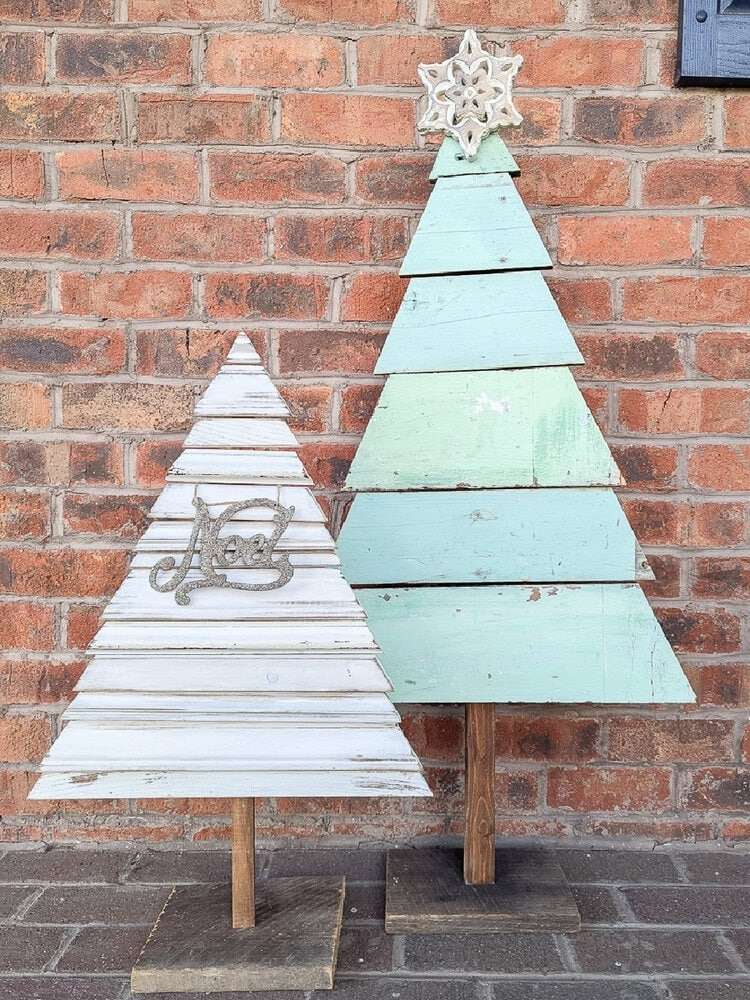 Two wooden outdoor Christmas trees, a smaller white one and a taller green one next to a brick wall
