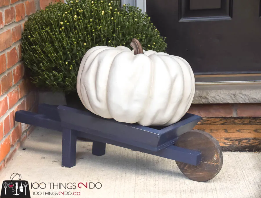 Dark blue painted wheelbarrow on a porch with a large white pumpkin and mum