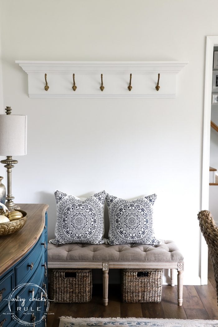 White coat rack with large hooks above a bench with pillows