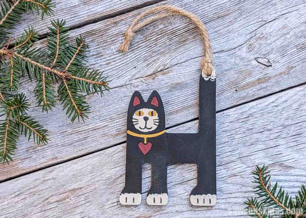 Front view of a folk art cat ornament on silver-colored wood with two sprigs of pine.