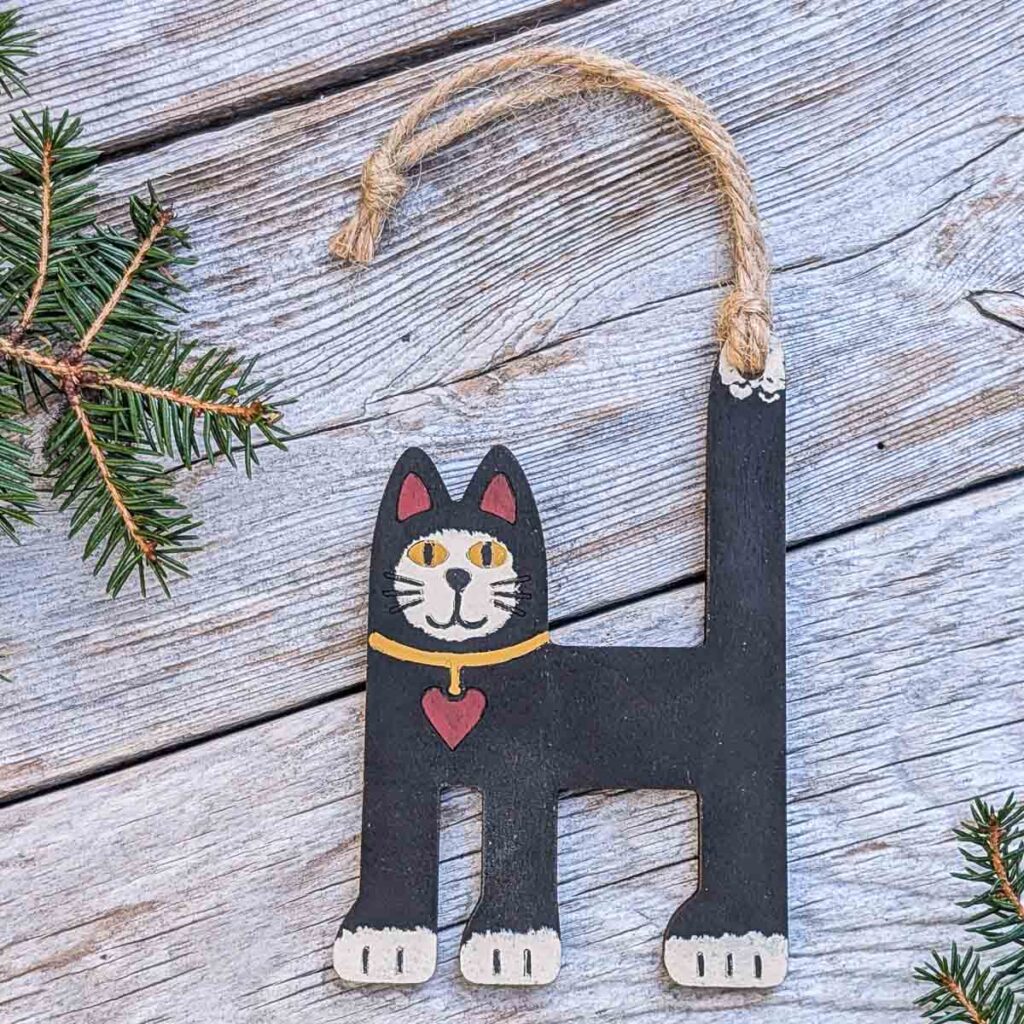 Front view of a folk art cat ornament on silver-colored wood with two sprigs of pine.