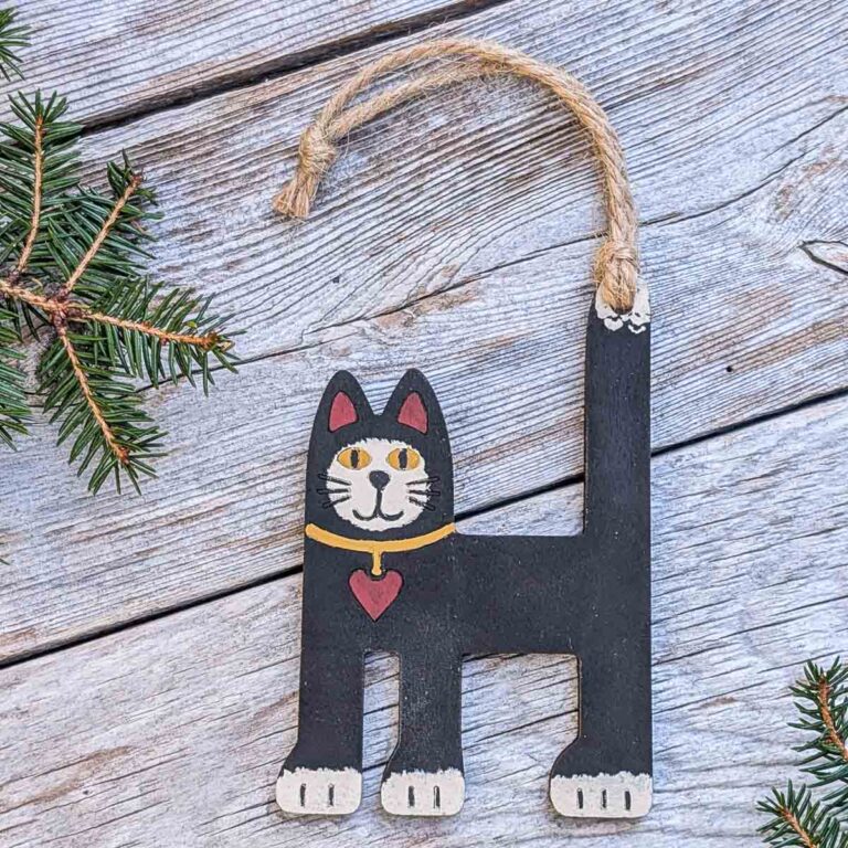 Front view of a folk art cat ornament on silver-colored wood with two sprigs of pine.
