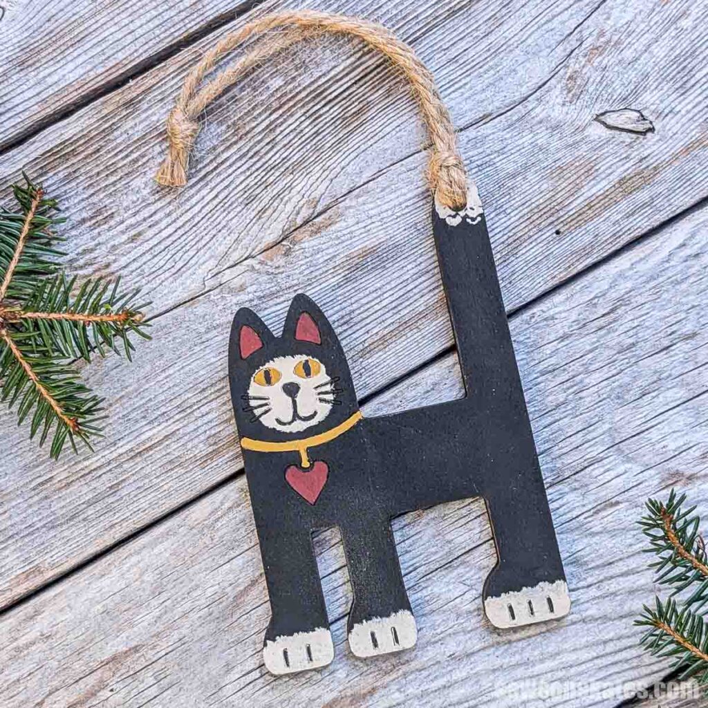 Angled view of a folk art cat ornament on silver-colored wood with two sprigs of pine.